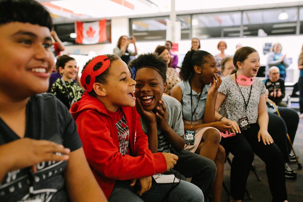 A photo of children sitting in a row laughing and showing excitement for what is happening