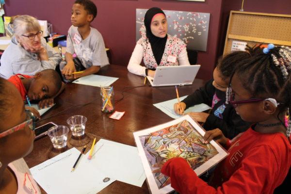 A woman in a hijab helps a group of children look at and write about a colorful abstract work of art