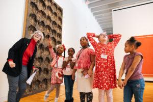 A group of five children stand in an art gallery with an adult. The children have looks of excitement on their faces